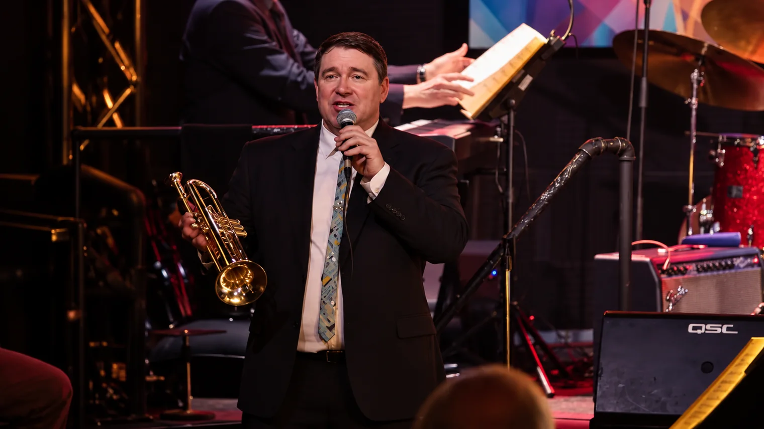 Chad Willis introducing music with microphone and trumpet — approved stage image illustrating his musical leadership