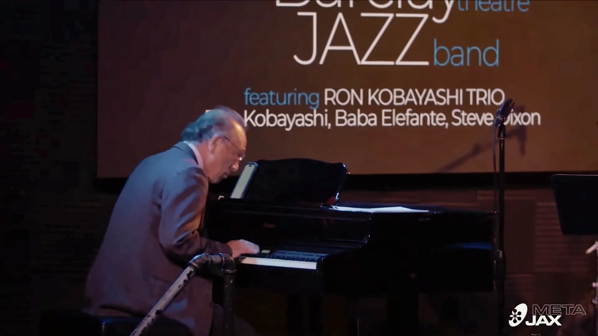 Ron Kobayashi at the grand piano with Ron Kobayashi Trio stage credits behind him